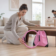 Woman kneeling at home and reaching for a pink resistance ring beside a pink drawstring bag, while another adult stands by a child in a cot in the background.